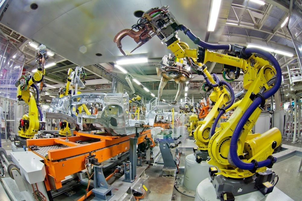 Heavy-duty industrial robotic arms, specifically bright yellow Fanuc robots, working on an automated automotive assembly line to weld and construct car body frames.